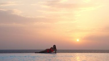 Mayolu ve güneş gözlüklü genç bir kadın güneş doğarken panoramik deniz manzaralı sonsuz bir havuzun kenarında uzanıyor ve dinleniyor. seyahat ve tatil kavramı