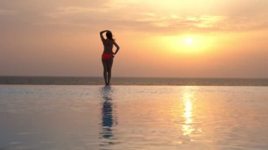 Arka manzara, mayo giymiş bir kadın silueti güneş doğarken panoramik deniz manzaralı açık hava sonsuz havuzunun kenarında duruyor ve dinleniyor. seyahat ve tatil kavramı