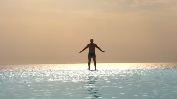Σκούρα σιλουέτα, ο άνθρωπος στέκεται με τα χέρια απλωμένα στις πλευρές και χαλαρώνοντας στην άκρη της εξωτερικής πισίνας απείρου με πανοραμική θέα στη θάλασσα, την ανατολή του ηλίου. έμπνευση, ταξίδια και διακοπές έννοια — Αρχείο Βίντεο