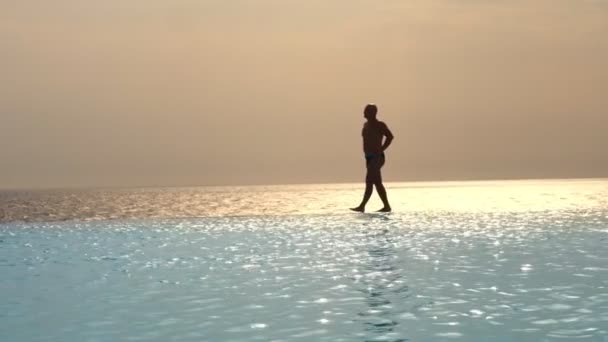 Silueta oscura, hombre caminando en el borde de la piscina infinita al aire libre con vistas panorámicas al mar, al amanecer. inspiración, viajes y concepto de vacaciones — Vídeo de stock