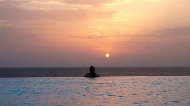 Vista para trás, silhueta de mulher, é relaxante na água, na borda da piscina exterior infinito com vista panorâmica do mar, ao nascer do sol. conceito de viagem e férias — Vídeo de Stock