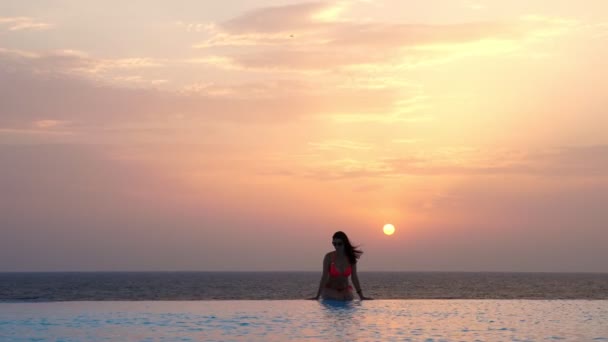 Молода жінка в купальнику і сонцезахисних окулярах, сидить і розслабляється на краю відкритого нескінченного басейну з панорамним видом на море, на сході сонця. концепція подорожей і відпусток — стокове відео