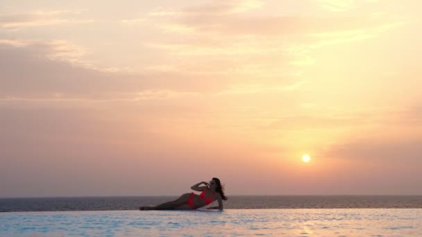 Jonge vrouw in badpak en zonnebril, ligt en ontspant aan de rand van het overloopzwembad met panoramisch uitzicht op zee, bij zonsopgang. reis- en vakantieconcept — Stockvideo