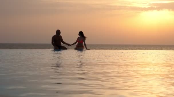 Silhouettes, couple in love, man and woman, sitting holding hands in outdoor infinity pool with panoramic sea view, at sunrise. honeymoon, travel and vacation concept — Stock Video