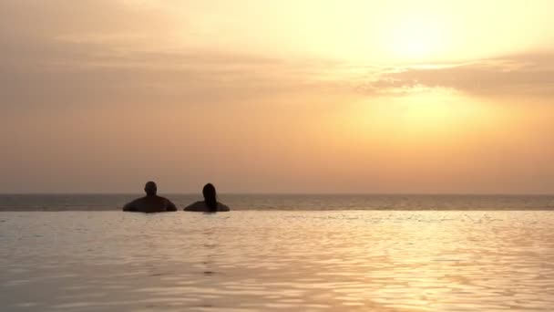 Pohled zezadu, siluety, zamilovaný pár, muž a žena, relaxace ve vodě, na okraji venkovního nekonečného bazénu s panoramatickým výhledem na moře, při východu slunce. Líbánky, cestování a dovolená koncept — Stock video