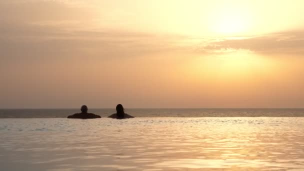 Back view, silhouettes, couple in love, man and woman, relaxing in water, on edge of outdoor infinity pool with panoramic sea view, at sunrise. honeymoon, travel and vacation concept — Stock Video