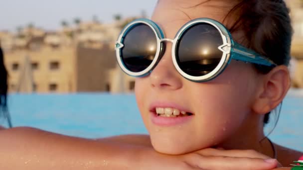 Retrato de uma adolescente sorridente, vestindo engraçado, óculos de sol redondos, relaxando na piscina exterior de um hotel elegante. O nascer do sol reflecte-se nos seus óculos. férias e viagens . — Vídeo de Stock