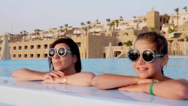 Donna e teen girl, mamma e figlia, in divertenti, identici, occhiali da sole rotondi, rilassarsi in acqua della piscina all'aperto con vista su un hotel di lusso con palme. concetto di vacanza familiare — Video Stock