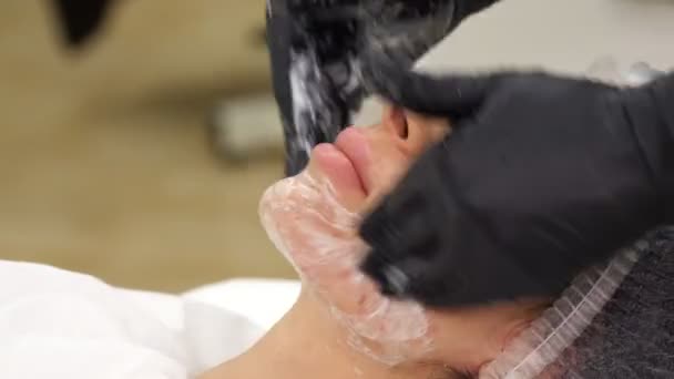 Close-up, beautician in black medical gloves performs cosmetic skincare procedure, applies cleansing foam or emulsion to female clients face. cosmetology, beauty salon. spa concept — Stock Video
