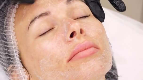 Närbild, kosmetolog i svarta medicinska handskar applicerar kosmetisk ansiktsskrubb med massagerörelser på kvinnligt ansikte. hudvård förfarande i kosmetologi klinik eller skönhetssalong — Stockvideo