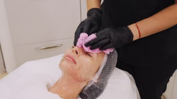 Zbliżenie, kosmetolog, w czarnych rękawiczkach medycznych, usuwa resztki kosmetycznego produktu do twarzy, z kobiecej twarzy za pomocą specjalnych chusteczek mokrych. zabieg pielęgnacji skóry w klinice kosmetologicznej lub salonie kosmetycznym. — Wideo stockowe