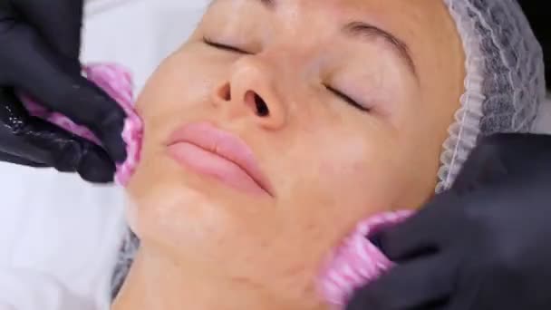 Zblízka kosmetolog v černých lékařských rukavicích odstraňuje zbytky kosmetického obličejového výrobku z ženského obličeje se speciálními mokrými ubrousky. péče o pleť v kosmetické klinice nebo kosmetickém salonu. — Stock video