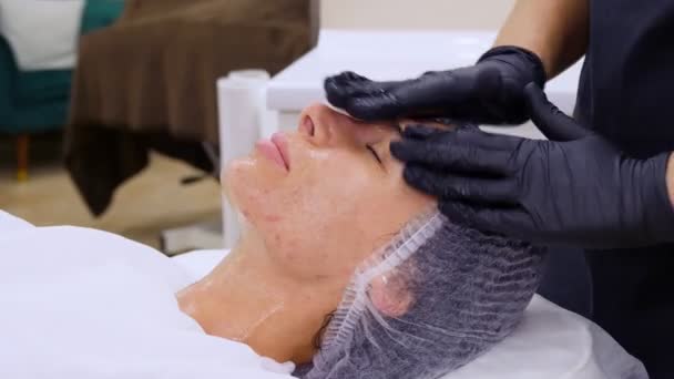 Närbild, kosmetolog i svarta medicinska handskar applicerar kosmetisk gel på kvinnligt ansikte med massagerörelser. hudvård förfarande i kosmetologi klinik eller skönhetssalong. — Stockvideo