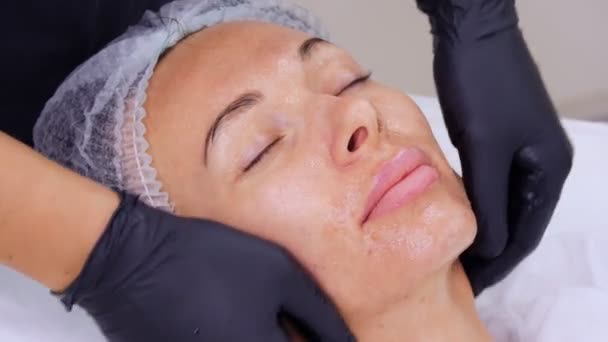 Primer plano, cosmetólogo, en guantes médicos negros, realiza el procedimiento de cuidado de la piel, masaje facial a una mujer, utilizando gel cosmético especial, en la clínica de cosmetología o salón de belleza. — Vídeo de stock
