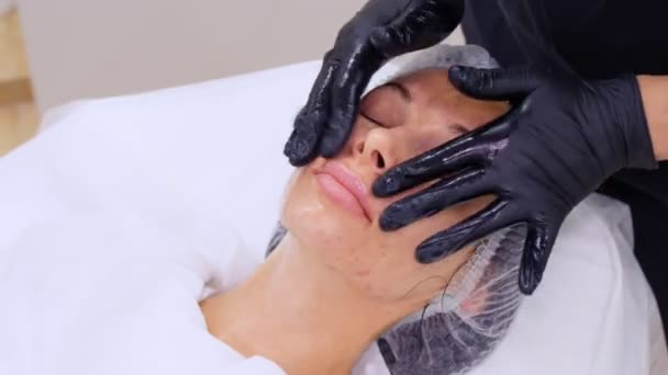 Närbild, kosmetolog, i svarta medicinska handskar, utför hudvård förfarande, ansiktsmassage till en kvinna, med hjälp av speciella kosmetiska gel, i kosmetologi klinik eller skönhetssalong. — Stockvideo