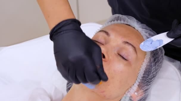 Close-up, κοσμητικός με μαύρα γάντια, εφαρμόζει μπλε καλλυντική μάσκα προσώπου με ειδικό πινέλο στο γυναικείο πρόσωπο. διαδικασία περιποίησης δέρματος σε κλινική κοσμετολογίας ή ινστιτούτο αισθητικής. — Αρχείο Βίντεο