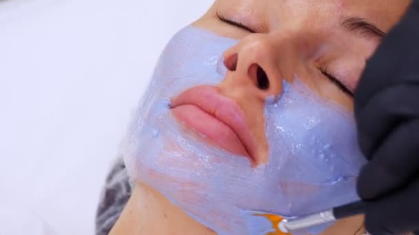 Primer plano, cosmético en guantes negros, aplica mascarilla cosmética azul con cepillo especial a la cara femenina. procedimiento de cuidado de la piel en la clínica de cosmetología o salón de belleza. — Vídeos de Stock