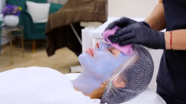 Крупним планом, косметолог, в чорних медичних рукавичках видаляє залишки косметичної маски для обличчя, з жіночого обличчя спеціальними мокрими серветками. процедура скінаре в косметологічній клініці або салоні краси . — стокове відео