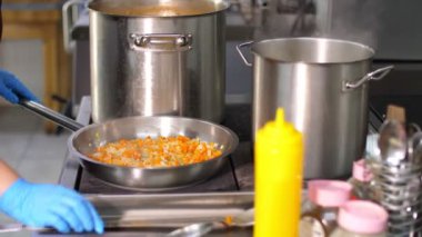 Kantinde yemek pişirmek. Yakın plan. Ocakta büyük tencereler var. Soğan ve havuç tavada kızartılır. Aşçı, koruyucu eldivenler içinde, kızartmayı karıştırıyor, ve sonra da çorba. sağlıklı gıda ve güvenlik kavramı