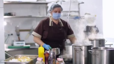 Yemek pişirmek. Kadın şef, koruyucu eldivenler ve maske takmış, mutfakta yemek hazırlıyor, kantinde. Sağlıklı gıda ve güvenlik kavramı. Matarayı yeniden aç