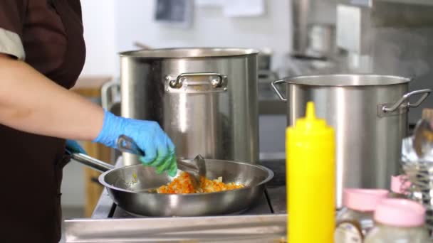 Cocinar en la cantina. Primer plano. hay ollas grandes en la estufa. La cebolla y la zanahoria se fríen en sartén. el cocinero, en guantes protectores, revolviendo la fritura. concepto de salud alimentaria y seguridad. reapertura — Vídeo de stock