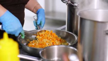 Yemek pişirmek. Yakın plan. Aşçı koruyucu eldivenlerle, kızartma tavasında kızarmış soğan ve havuçları karıştırıyor. Sağlıklı gıda ve güvenlik kavramı. Matarayı yeniden aç