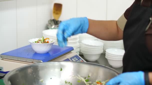 Koken. close-up. de kok doet in beschermende handschoenen groentesalade in kleine schaaltjes en weegt deze op een weegschaal. Gezondheidsvoedsel. vrijwilligerswerk en liefdadigheid. Kantine heropenen. veiligheidsconcept. — Stockvideo