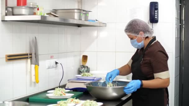 Cucinare. La cuoca, in maschera protettiva e guanti, mette l'insalata vegetale in piccola ciotola e la pesa su una scala. cibo salutare. volontariato e carità. Riaprire la mensa. concetto di sicurezza. — Video Stock