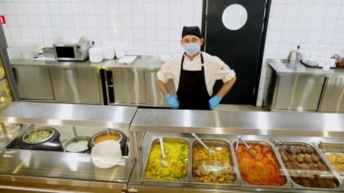 Koruyucu eldiven ve maske takmış bir aşçı ya da garson farklı yemeklerle sergi masasının arkasında durur, self-servis kafeteryasında ya da açık büfede. Sağlıklı yemek. Gönüllü olmak ve yardım. yeniden açılıyor