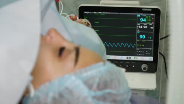 Primer plano. paciente mujer, en estado de anestesia, se encuentra en la mesa de operaciones, frente al monitor, que muestra la presión arterial del paciente, el ritmo y los latidos del corazón, durante la cirugía — Vídeo de stock