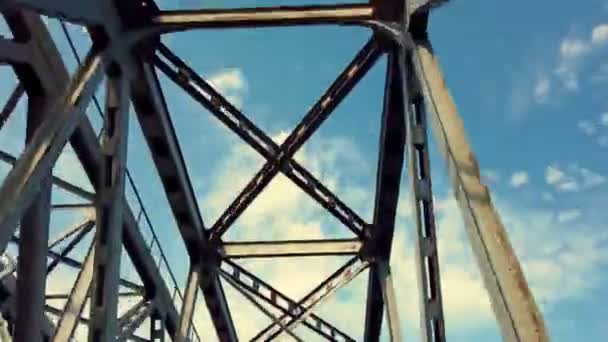 Eisenkonstruktion der Brücke gegen blauen Himmel mit weißen Wolken. Fahrt unter Brücke, Blick auf Brücke von unten. Silhouette von kreuzenden Stahlträgern. Brückendetails gegen strahlend blauen Himmel. — Stockvideo