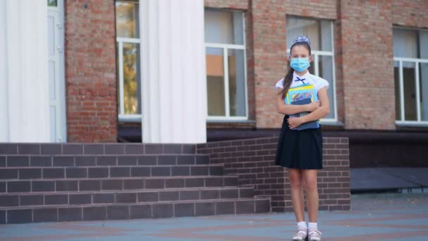 Al aire libre. colegiala, adolescente, en máscara de protección médica, con libros en sus manos, en el fondo del edificio de la escuela. Regreso a la escuela después del brote de covid19. — Vídeos de Stock