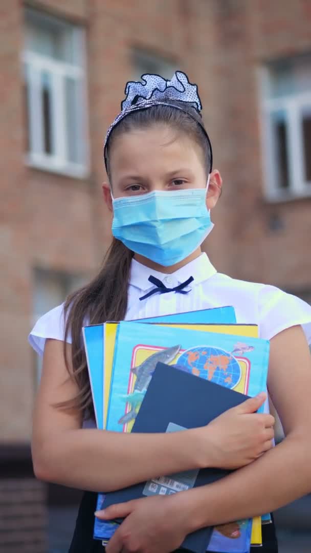 Verticale video. buitenportret van schoolmeisje, met medisch beschermend masker, met boeken in haar handen, op de achtergrond van school. Terug naar school na de uitbraak van de covid19. — Stockvideo