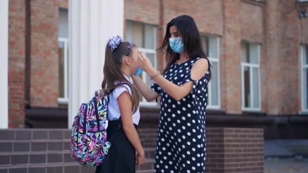 Mamá pone máscara protectora y médica en la cara de sus hijas colegialas y le da un pequeño aerosol antiséptico, sobre el fondo del edificio de la escuela. Regreso a la escuela después del brote de covid19. — Vídeos de Stock