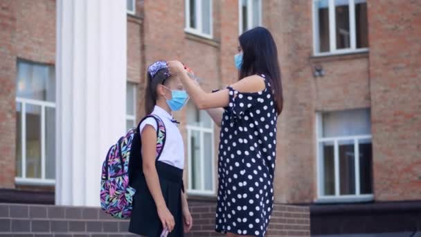 Maman met un écran protecteur sur sa fille écolière, sur le fond du bâtiment de l'école. Retour à l'école après l'éclosion de covid19. — Video