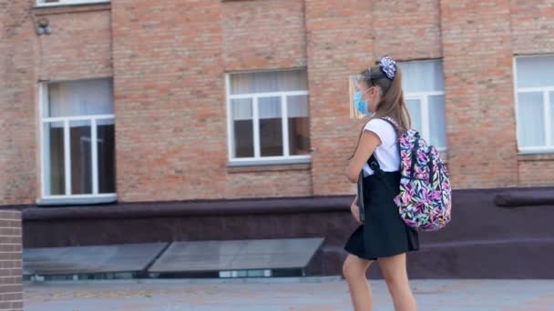 Al aire libre, colegiala, en máscara protectora y escudo, con mochila, va a la escuela a través del patio de la escuela. Regreso a la escuela después del brote de covid19. — Vídeos de Stock