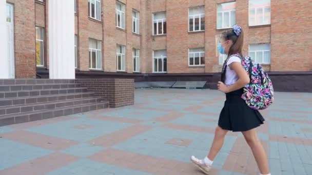 Utomhus, skolflicka, i skyddsmask och sköld, med ryggsäck, går i skolan över skolgården. Tillbaka till skolan efter covid19 utbrott. — Stockvideo