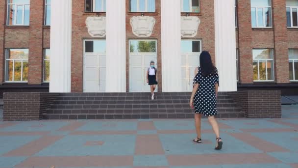 Mamá conoce a su hija de la escuela en el patio de la escuela. colegiala y mujer, ambos están en máscaras protectoras. Regreso a la escuela después del brote de covid19. Saludos en Covid-19 tiempo — Vídeos de Stock