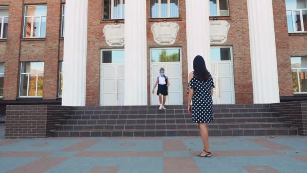 Mamá conoce a su hija de la escuela en el patio de la escuela. colegiala y mujer, ambos están en máscaras protectoras. Regreso a la escuela después del brote de covid19. Saludos en Covid-19 tiempo — Vídeos de Stock