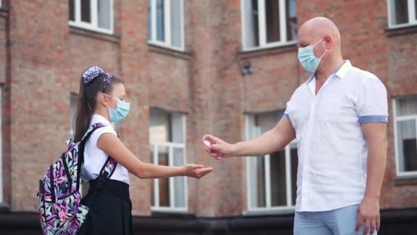マスクの男は女子学生の手に防腐剤を振りかけ校舎を背景に。covid19発生後に学校に戻る. — ストック動画