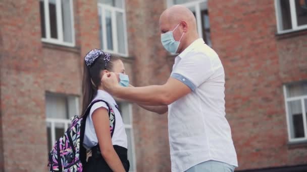 Pappa sätter skyddande, medicinsk ansiktsmask på sin dotter skolflicka och ger små antiseptiska spray, på skolan byggnad bakgrund. Tillbaka till skolan efter covid19 utbrott. — Stockvideo