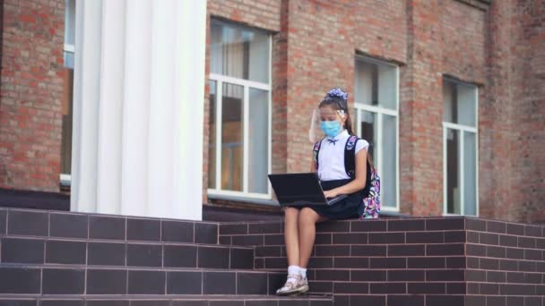 Skolflicka, tonåring flicka, i medicinsk mask och skyddande ansiktsskydd, använder laptop, medan du sitter på skolgården. Tillbaka till skolan efter covid19 utbrott. — Stockvideo