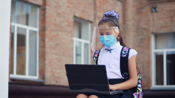 Schoolmeisje, tienermeisje, met medisch masker en beschermend gezichtsschild, gebruikt laptop, terwijl ze op het schoolplein zit. Terug naar school na de uitbraak van de covid19. — Stockvideo