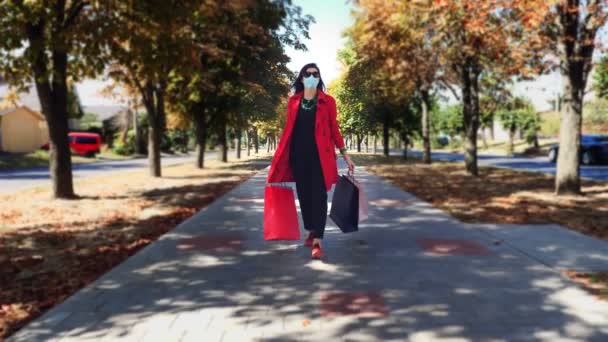 Mujer con bolsas de compras. mujer, en máscara protectora, con bolsas de compras de colores en sus manos, caminando por el callejón de la ciudad. compras y regalos. concepto de entrega o donación. — Vídeos de Stock