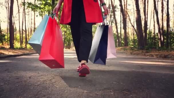 Vrouw met boodschappentassen. Vrouwelijke benen in rode schoenen, close-up. Vrouw met gekleurde boodschappentassen in haar handen, lopend door het stadspark. winkelen en cadeaus. leverings- of donatieconcept. — Stockvideo