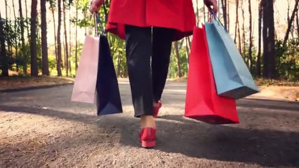 Kvinna med shoppingväskor. Honben i röda skor, närbild. Kvinna med färgade shoppingväskor i händerna, promenader genom stadsparken. shopping och presenter. leverans- eller donationskoncept. — Stockvideo