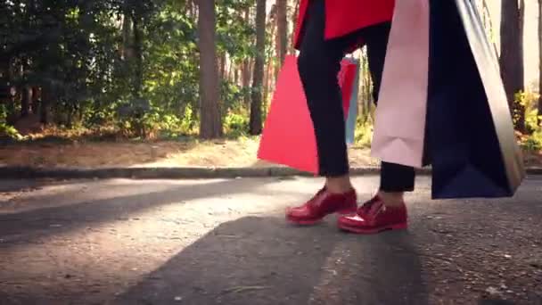 Femme avec des sacs à provisions. jambes féminines en chaussures rouges, gros plan. Femme avec des sacs à provisions colorés dans ses mains, marchant dans le parc de la ville. achats et cadeaux. concept de livraison ou de don. — Video