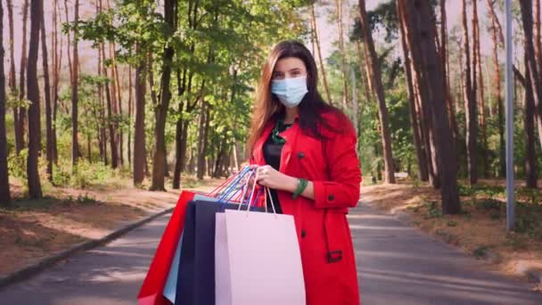 Egy nő bevásárlószatyorral. portré egy nőről, védőmaszkban, színes bevásárlószatyrokkal a kezében, a városi parkban. vásárlás és ajándékok. szállítási vagy adományozási koncepció. — Stock videók