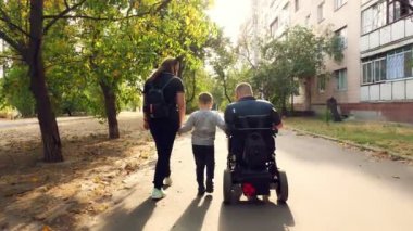Tekerlekli sandalye. Engelli adam. Otomatik tekerlekli sandalyeli genç bir adam güneşli bir sonbahar gününde ailesi, karısı ve küçük çocuğuyla birlikte şehir sokaklarında yürüyor. arka plan