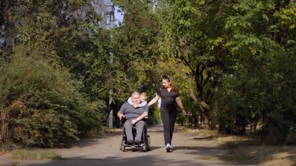 Wózek inwalidzki. Niepełnosprawny. młody niepełnosprawny mężczyzna na zautomatyzowanym wózku spacerowym z rodziną, żoną i małym dzieckiem, wzdłuż uliczki miejskiej w słoneczny jesienny dzień. — Wideo stockowe